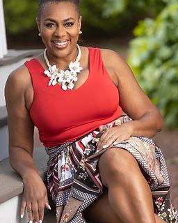Dr. Angie Kearns seated on wooden stairs outdoors, wearing a vibrant red V-neck top and a patterned floral skirt with a white floral statement necklace, warm direct smile, soft green foliage bokeh behind her.