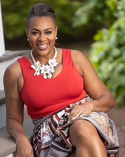 Dr. Angie Kearns seated on wooden stairs outdoors, wearing a vibrant red V-neck top and a patterned floral skirt with a white floral statement necklace, warm direct smile, soft green foliage bokeh behind her.