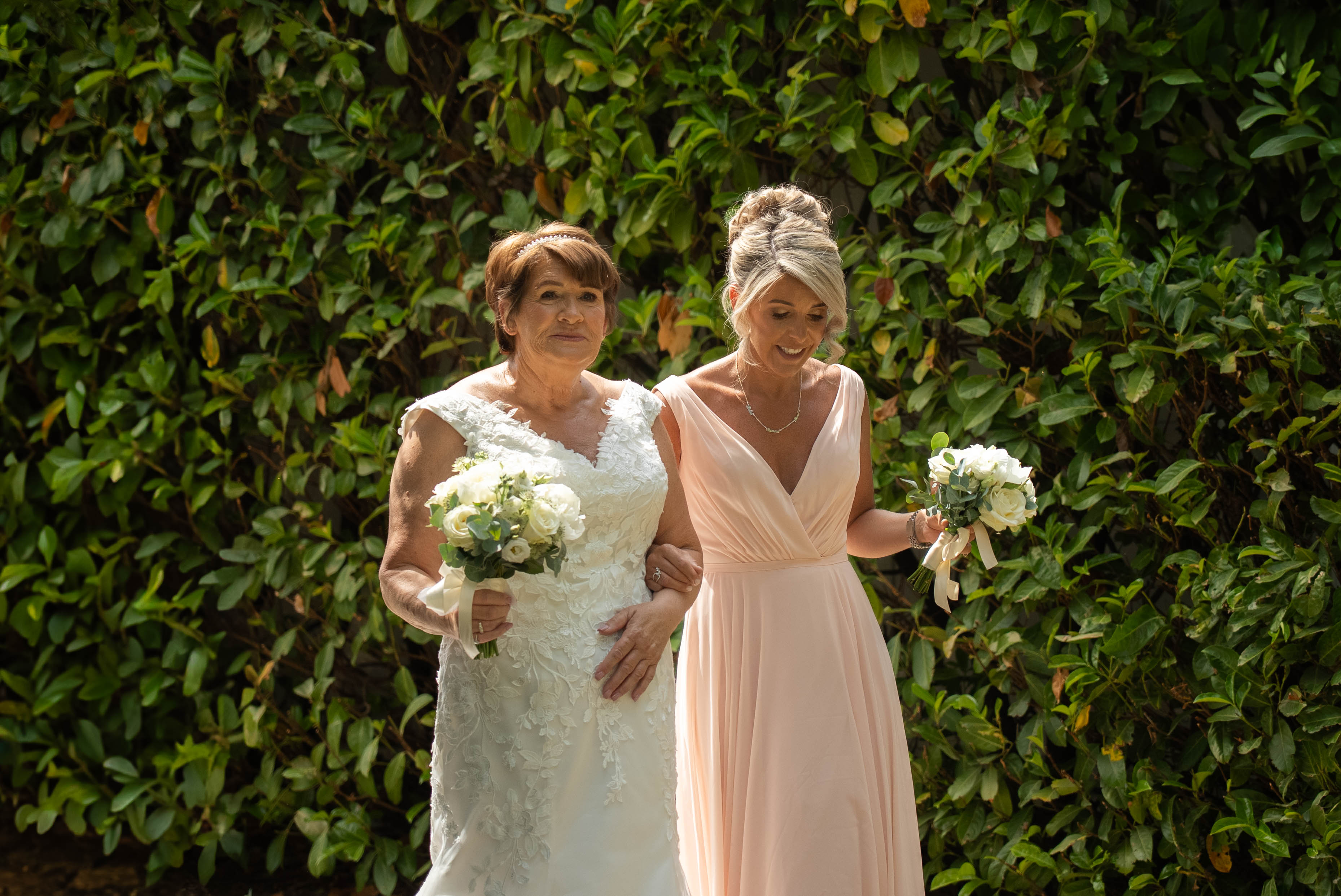 Bride and bridesmaid walking together in front of a green hedge