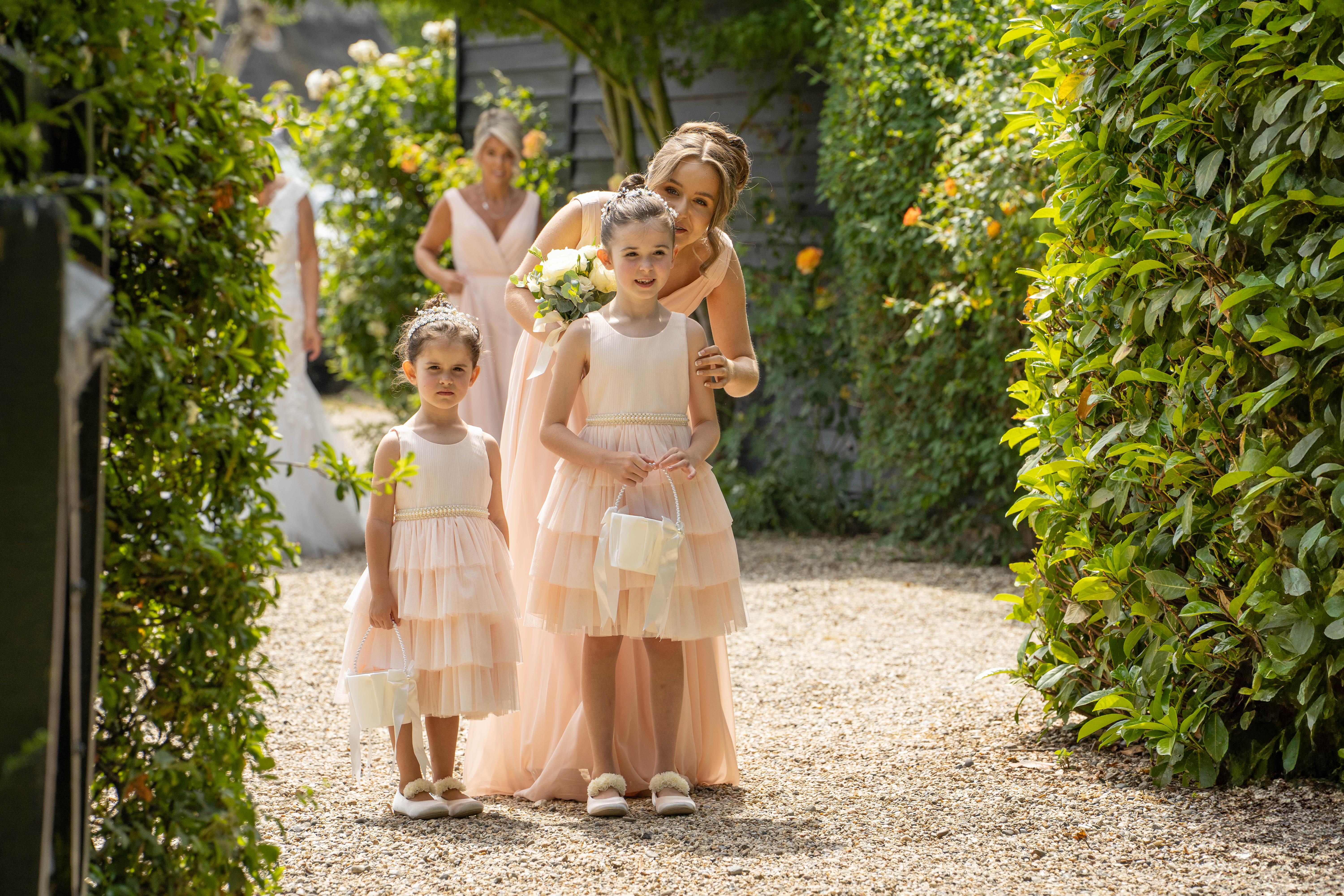 Flower girls in pale peach dresses standing on a gravel path lined with green hedges