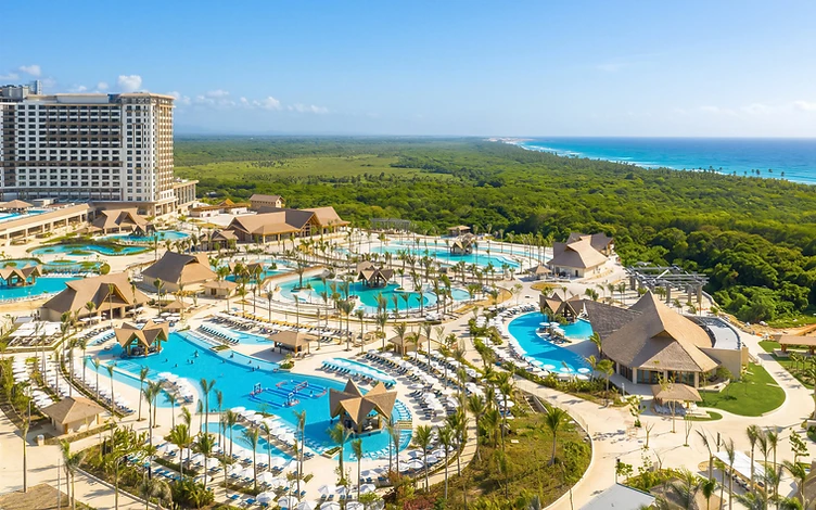 Aerial view of resort pools, palm-lined terraces and pitched-roof pavilions, with coastal greenery and the ocean beyond.