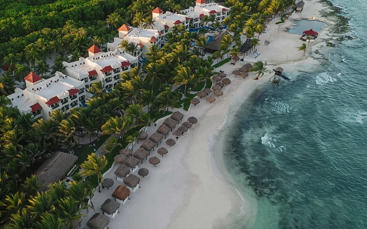 Aerial view of El Dorado Royale, with white resort buildings, palm trees and thatched shelters along a sandy beach.