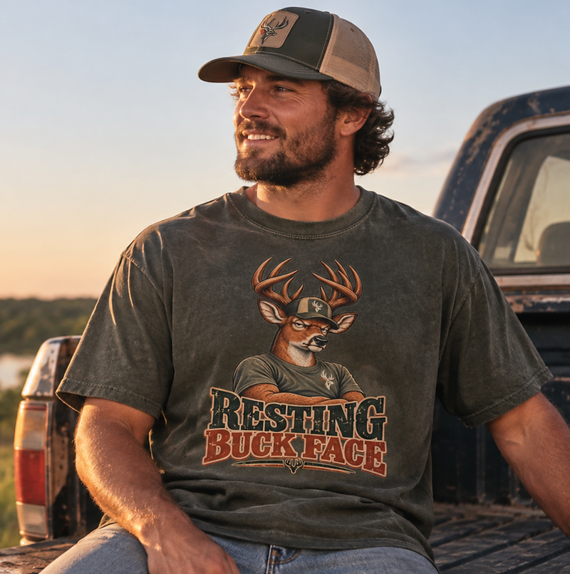 Man sitting on a pickup tailgate at sunset wearing a Lazy Buck Resting Buck Face shirt.