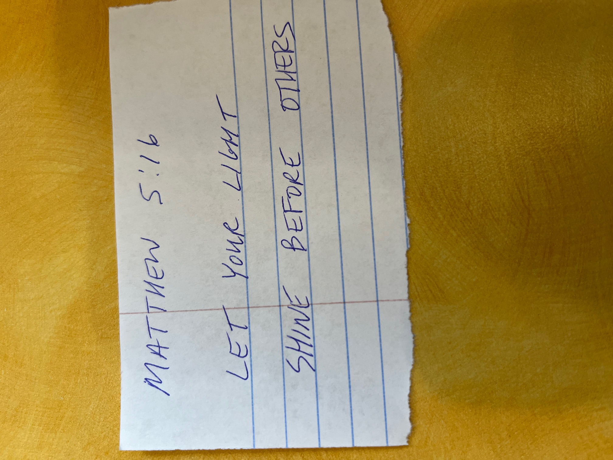 A torn piece of notebook paper with Matthew 5:16 handwritten in blue ink