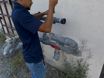 Plumber using a hand tool on exterior gas piping and a regulator mounted to a building's outside wall.