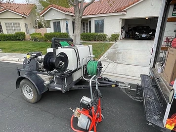 Service truck and jetting trailer with a water tank and hose reels parked on a residential street, with a drain-cleaning machine alongside.