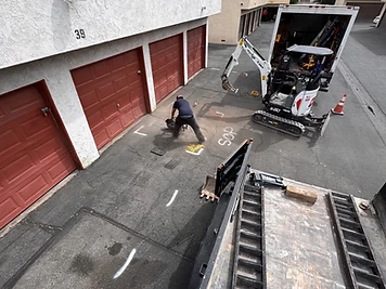 Plumber saw-cutting marked asphalt to open an excavation for a sewer-line repair, with a compact excavator nearby.