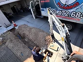 Plumber working in a trench at a water-line repair, with a compact excavator and service truck on a residential driveway.
