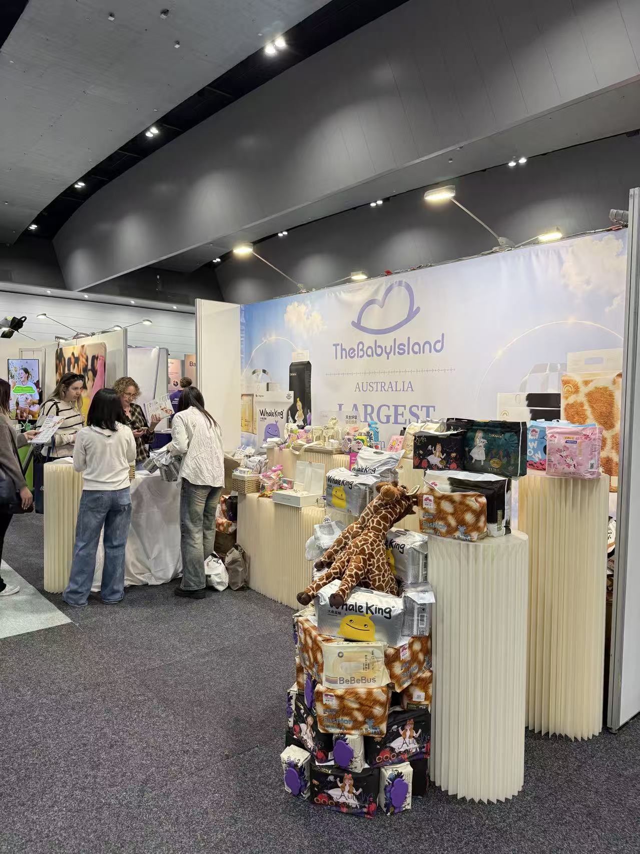 TheBabyIsland expo booth with product displays and plush giraffe