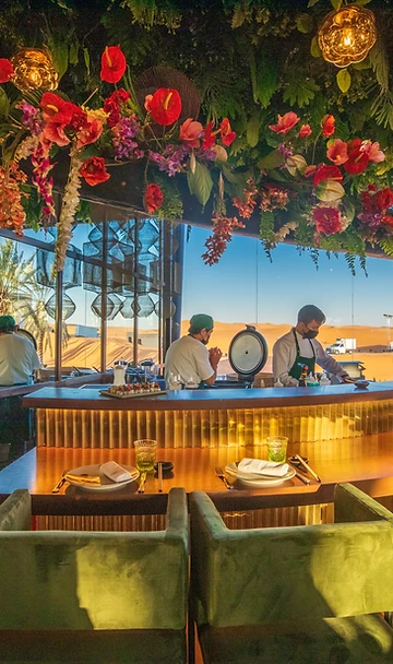 Luxury desert café interior with green velvet seating and floral ceiling installation overlooking dunes