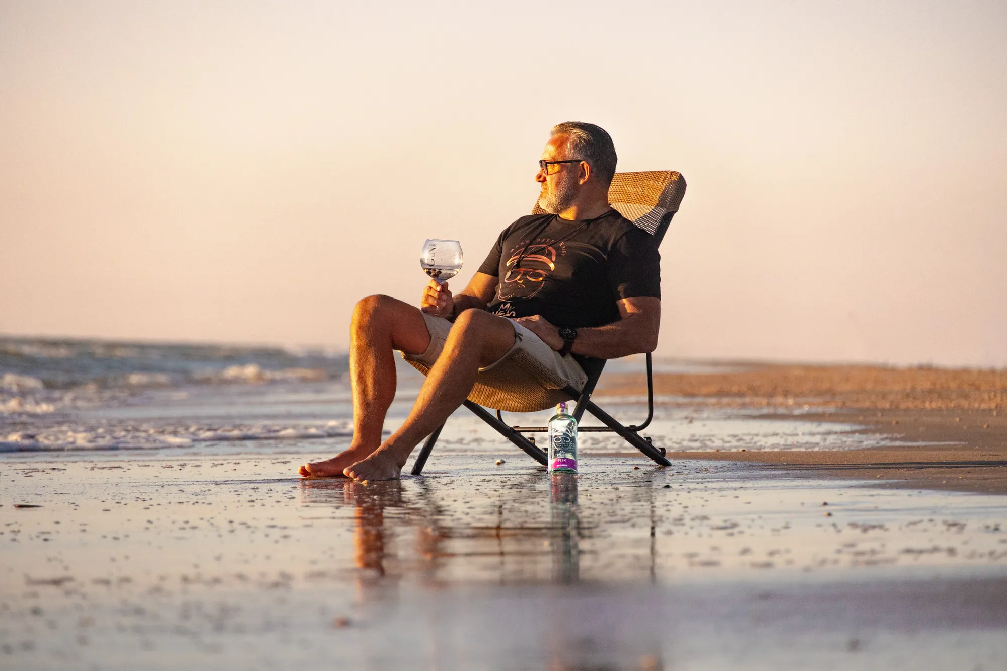 Mand i My Hero-t-shirt sidder på stranden ved solnedgang med et glas gin og en flaske foran sig