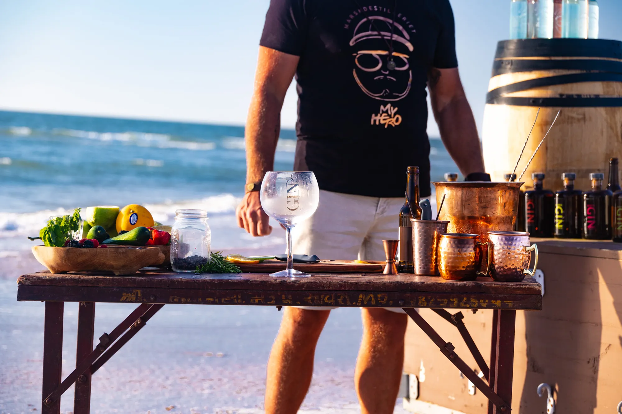 Person i My Hero-t-shirt forbereder gin-drinks ved et træbord på stranden