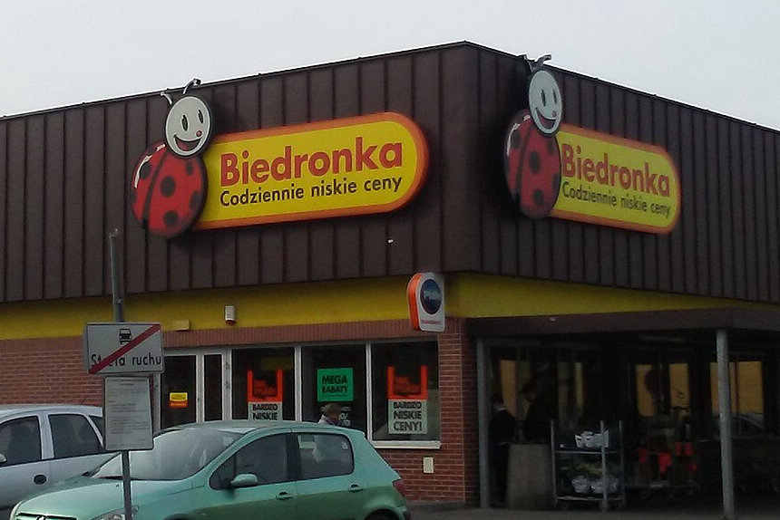 Sklep Biedronka z logo biedronki na brunatnej elewacji, przed budynkiem parking z kilkoma samochodami