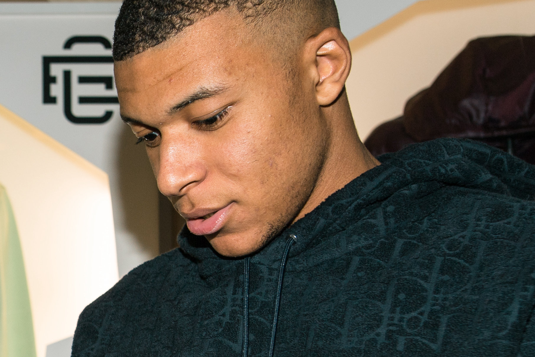 Kylian Mbappé w ciemnej bluzie z kapturem, z pochyloną głową, na tle ekspozycji odzieży