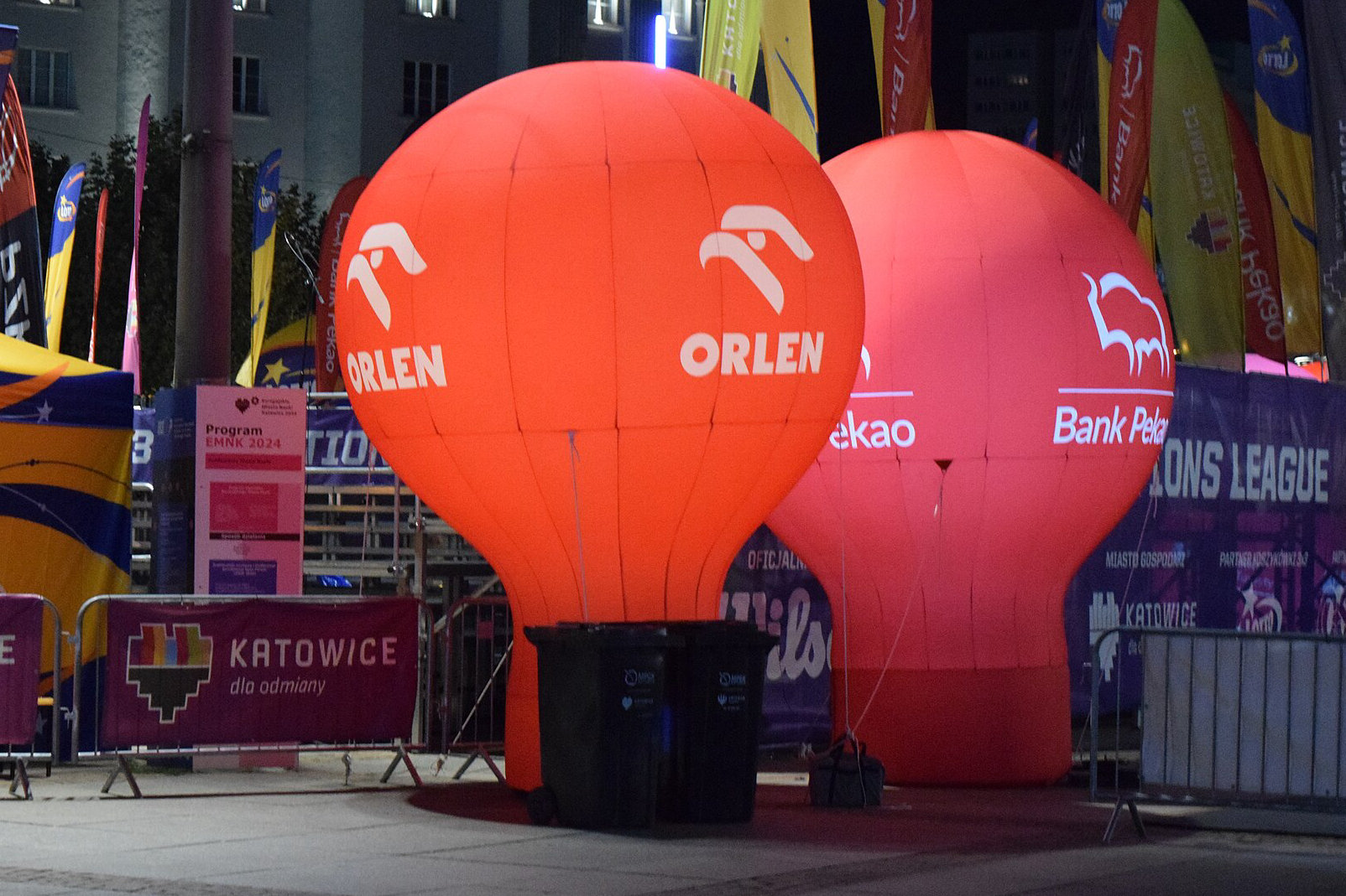 Nocą na katowickim rynku stoją dwa podświetlone czerwone balony reklamowe z logo Orlen i Banku Pekao, w tle flagi sponsorów wydarzenia.