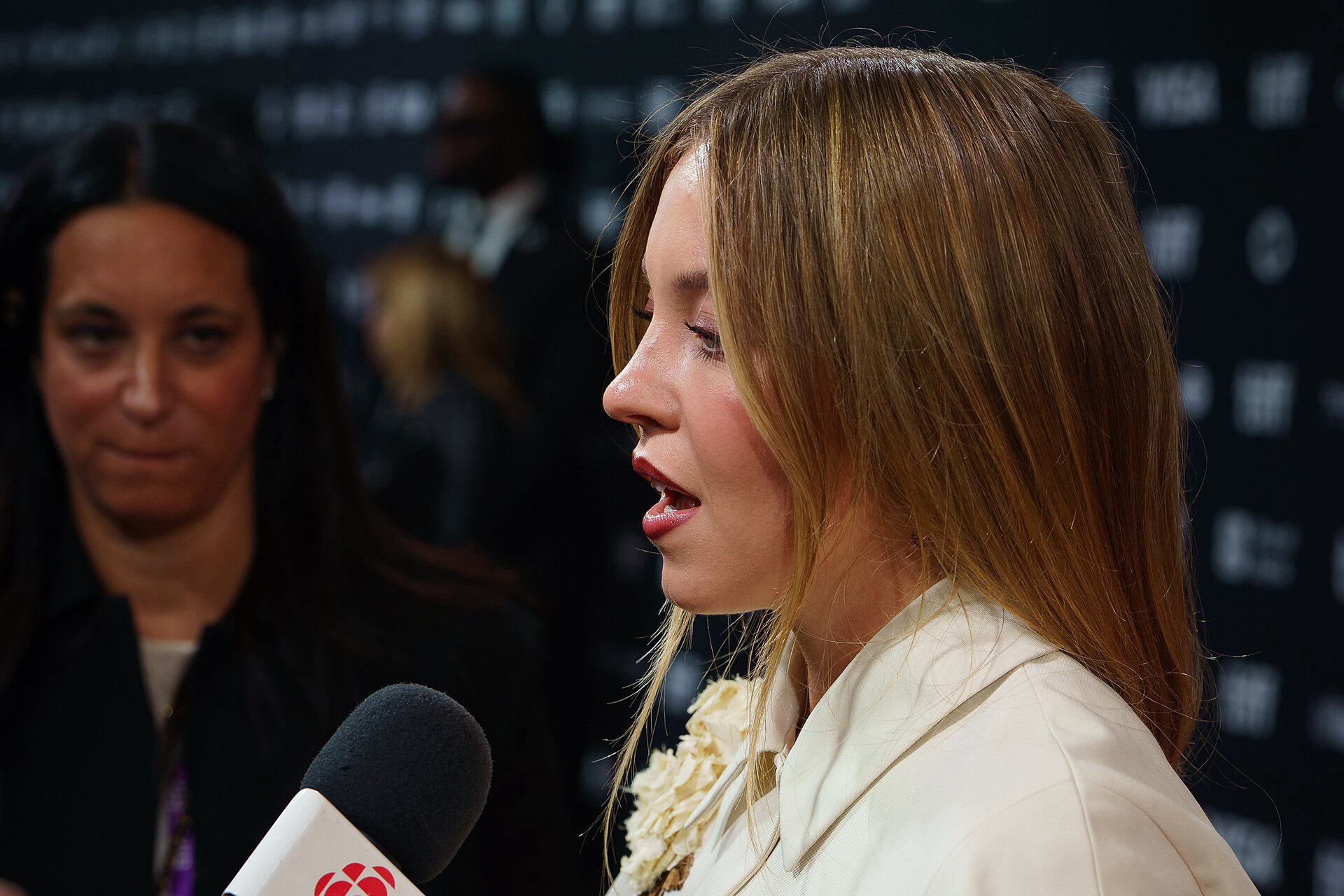Sydney Sweeney w kremowej stylizacji udziela wywiadu na czerwonym dywanie festiwalu TIFF 2024, przed ścianką z logotypami.