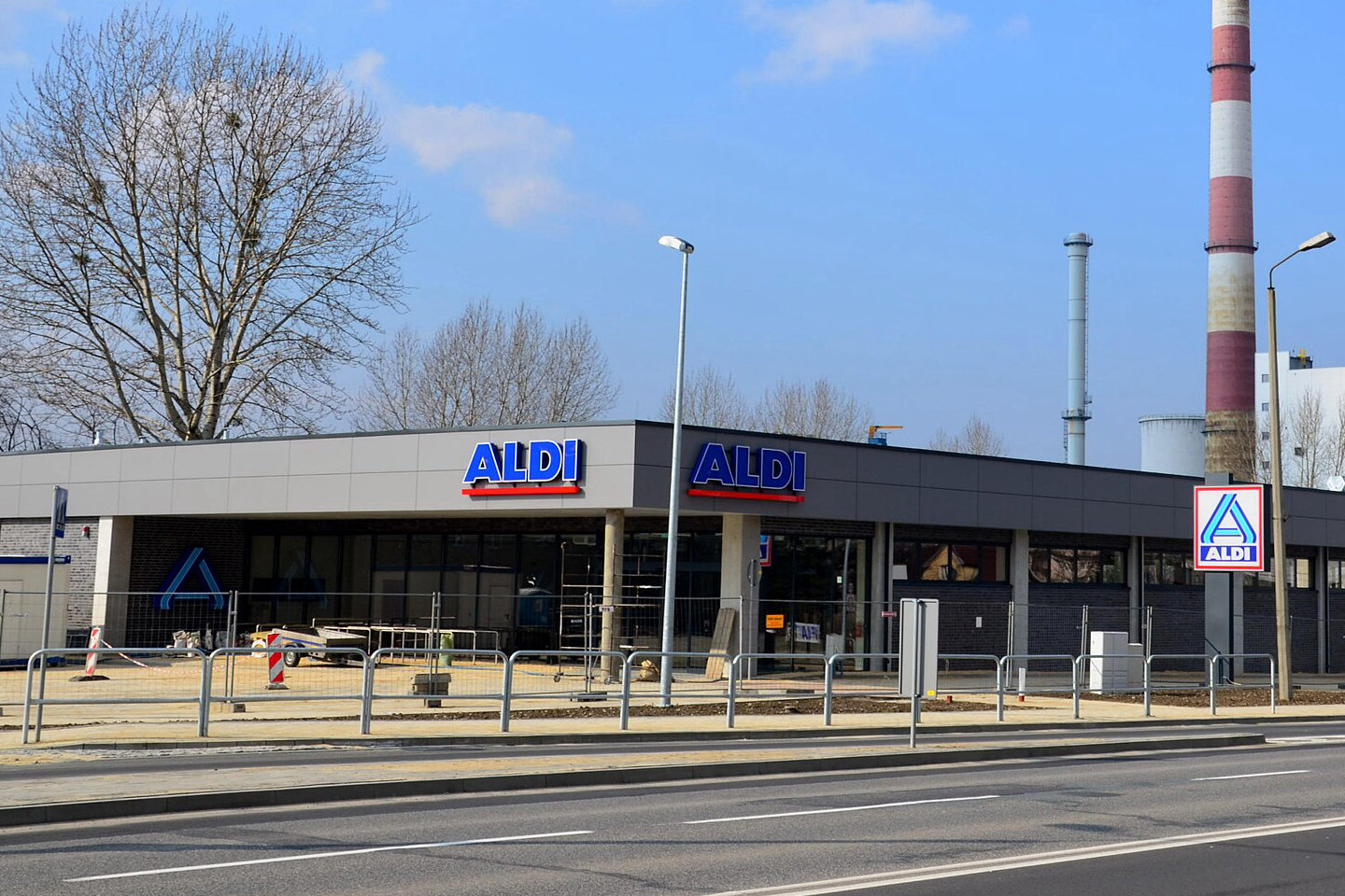Nowy sklep Aldi w Polsce z niebiesko-czerwonymi napisami ALDI na fasadzie i pylonem z logo, w tle kominy elektrociepłowni