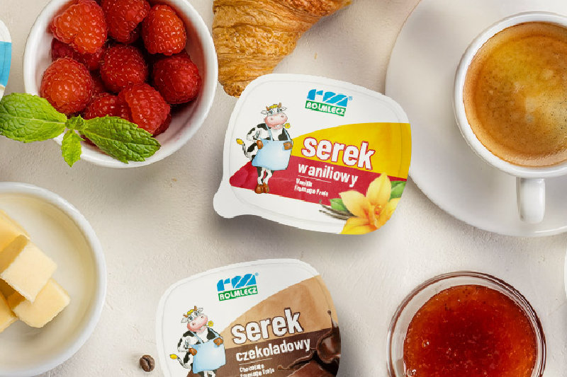 Opakowania serków Rolmlecz w różnych smakach na jasnym blacie wśród rogalików, malin, kawy, masła i dżemu