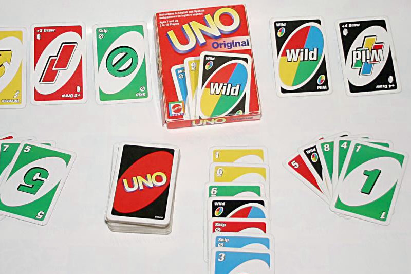 Karty do gry UNO Original rozłożone na białym tle, obok czerwone pudełko z logo Mattel oraz karty specjalne i talie graczy