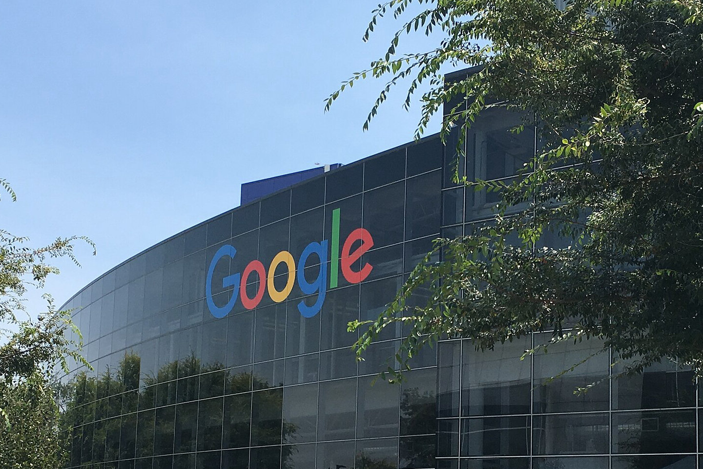 Szklana fasada siedziby Google w Mountain View z kolorowym logo, przed budynkiem trawa i drzewa
