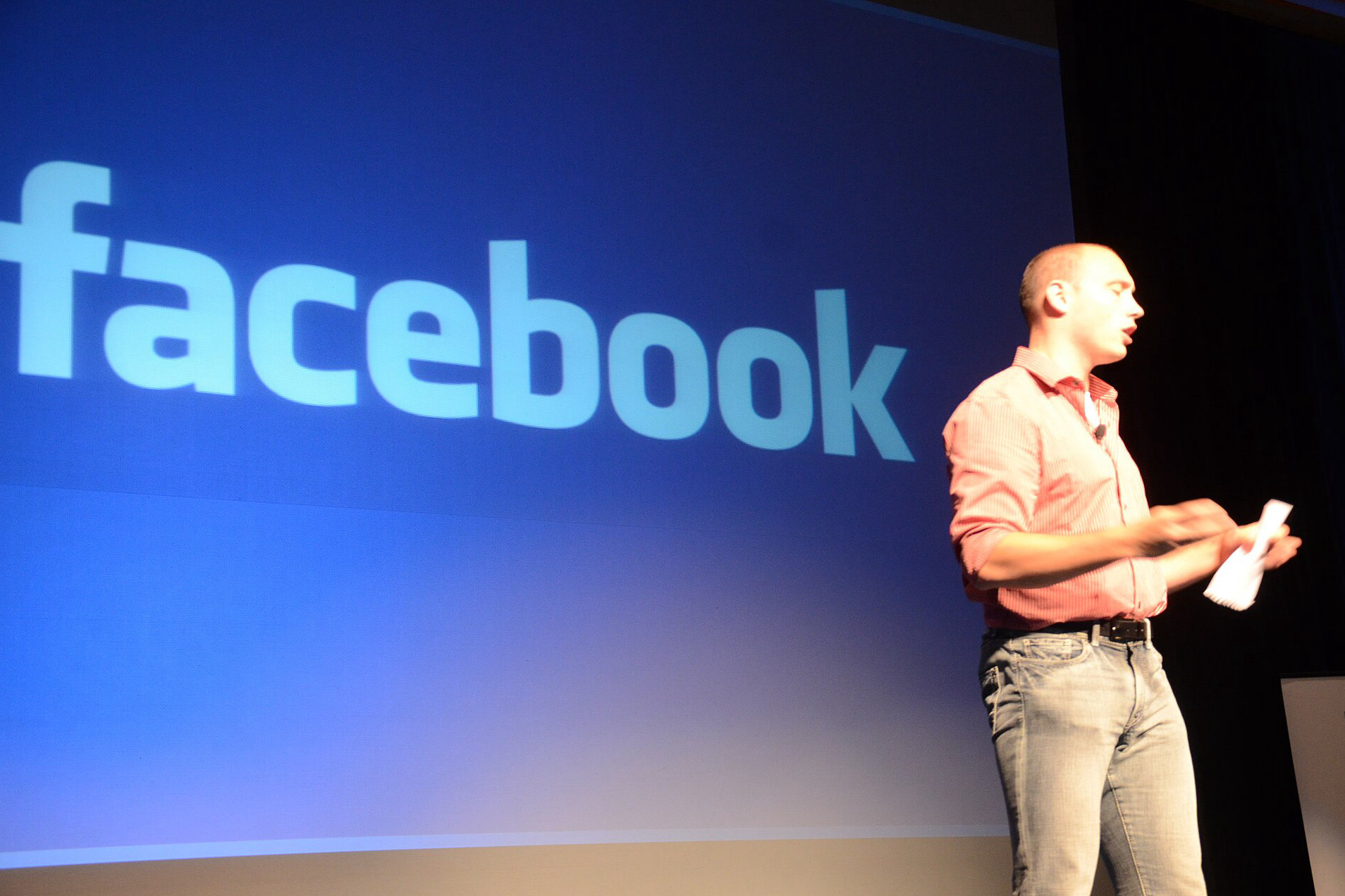 Mężczyzna w różowej koszuli przemawia na scenie na tle dużego niebieskiego ekranu z logo Facebooka