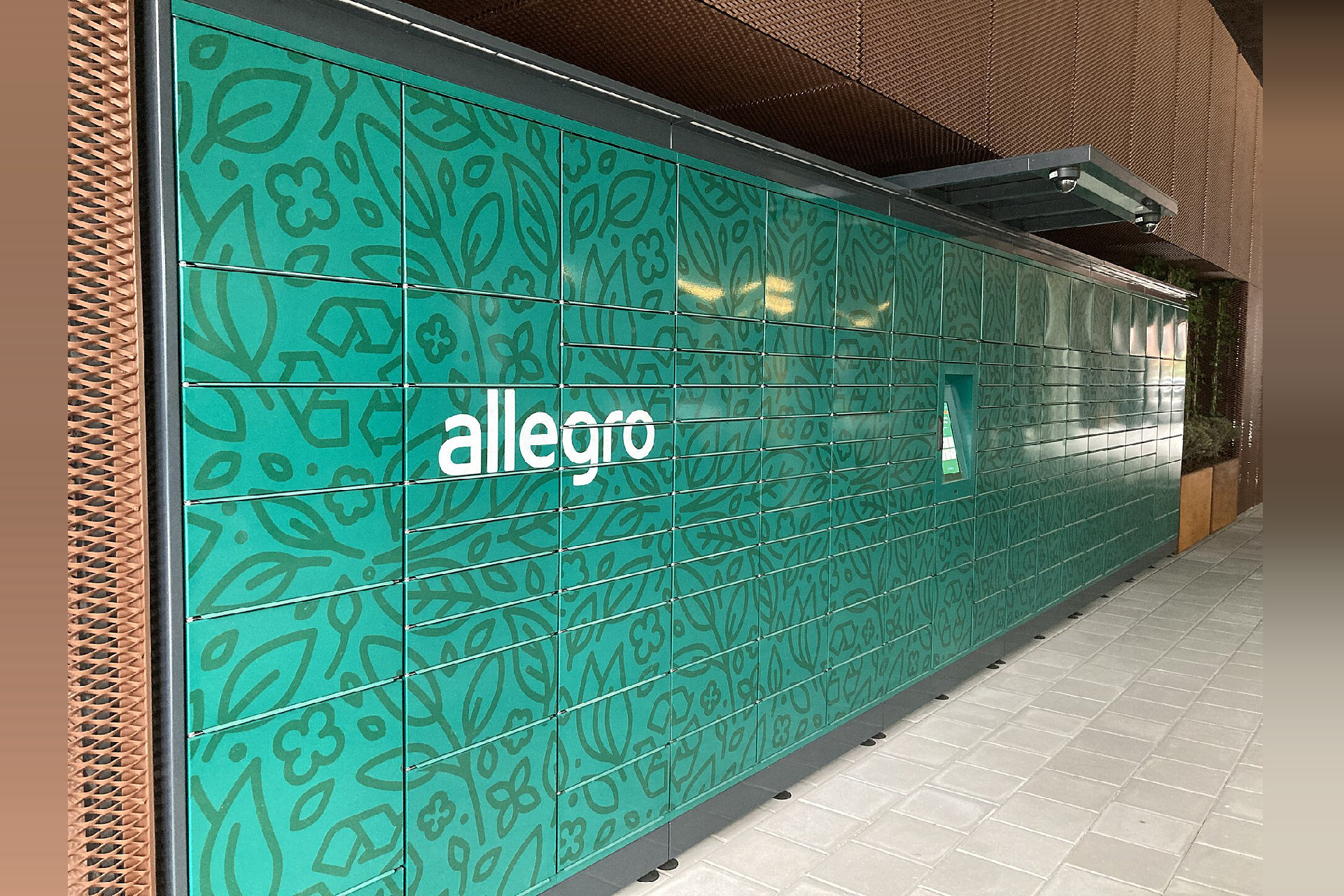 Zielony automat paczkowy Allegro z białym logo marki, ustawiony wzdłuż ściany budynku przy chodniku