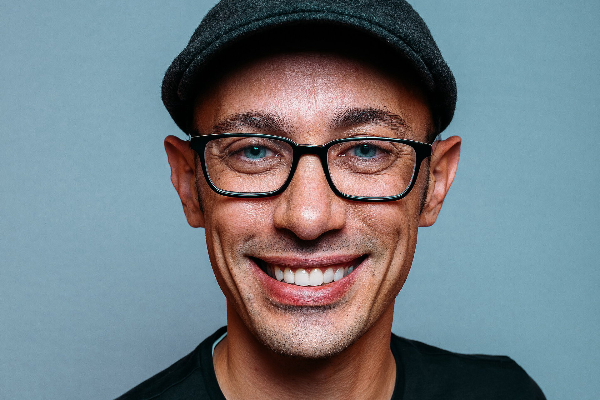 Tobias Lütke, szef Shopify, w czapce i okularach, uśmiechnięty, na jednolitym niebieskoszarym tle, portret z wydarzenia Shopify+ w 2019 roku