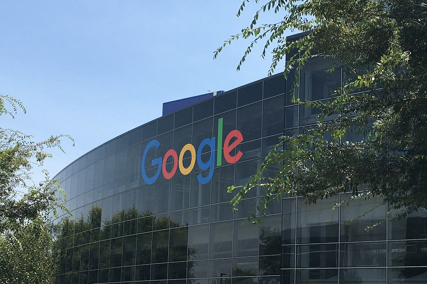 Szklana fasada budynku Googleplex w Mountain View z kolorowym logo Google, na pierwszym planie trawy i drzewa