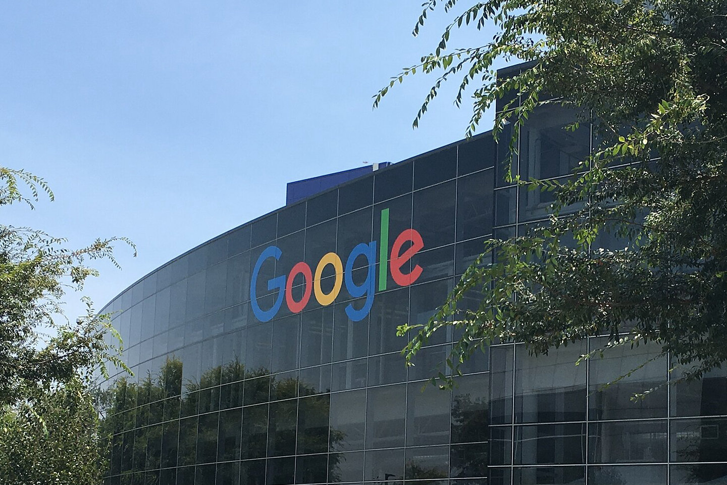 Przeszklona fasada siedziby Google w Mountain View z kolorowym logo Google, na pierwszym planie trawy i drzewa