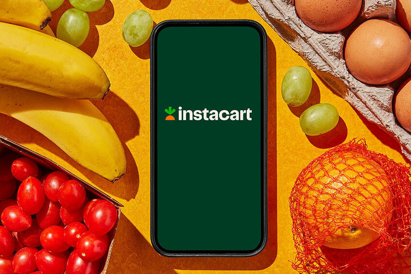 Smartfon z logo Instacart na żółtym tle, wokół banany, winogrona, pomidorki koktajlowe, jajka w tekturowej tacce i pomarańcza w siatce.