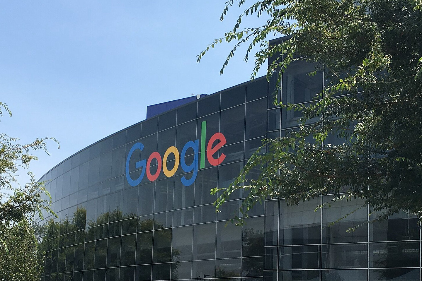 Przeszklona fasada siedziby Google w Mountain View z kolorowym logo firmy, w otoczeniu drzew i traw