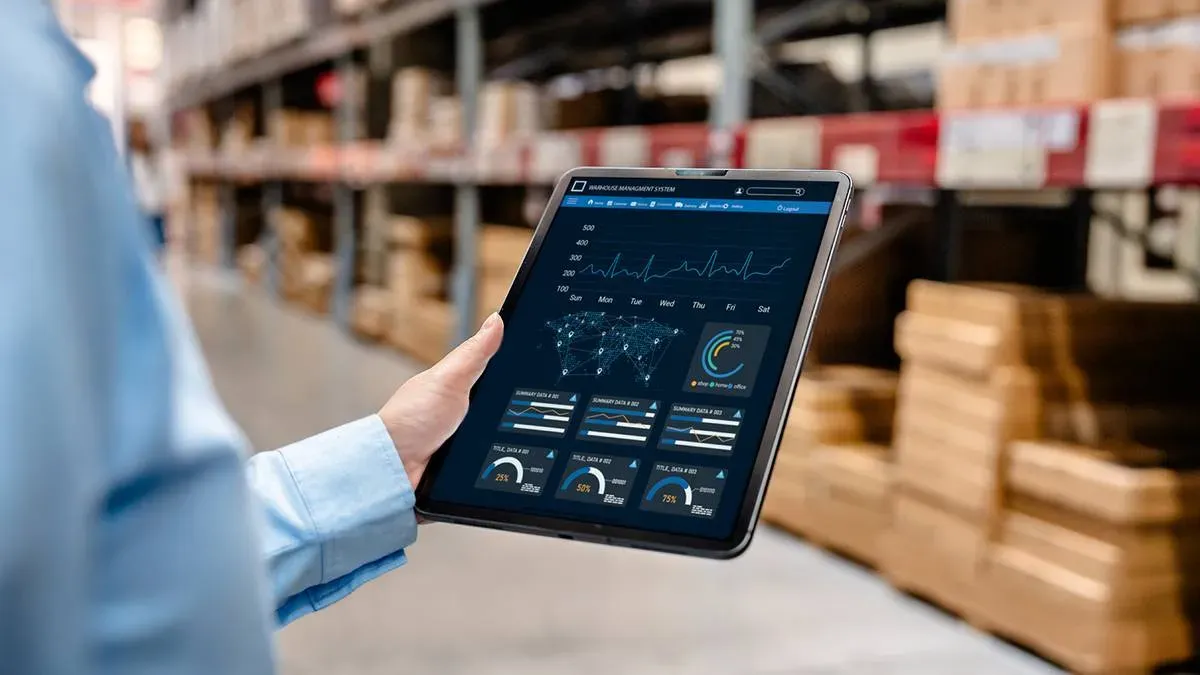 Dłoń trzymająca tablet z wykresami i danymi sprzedażowymi na tle regałów magazynowych z kartonami