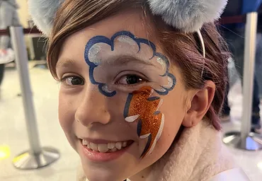 Smiling child with cloud and lightning bolt face paint and pom-pom accessories
