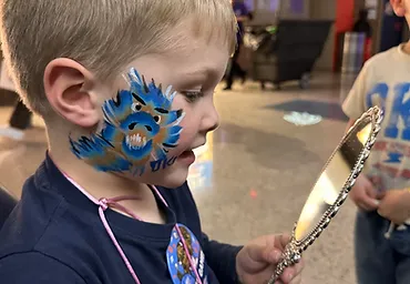 Boy admiring a blue and orange dragon face paint design in a hand mirror