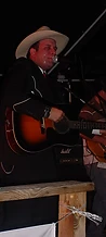 Joey Allcorn in a cowboy hat and suit jacket playing acoustic guitar on the night stage at JescoFest 2006