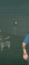 Bearded cowboy by a campfire while a man plays acoustic guitar at night at JescoFest 2006