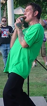 Jesco White playing harmonica on the grass while a woman photographs him at JescoFest 2006