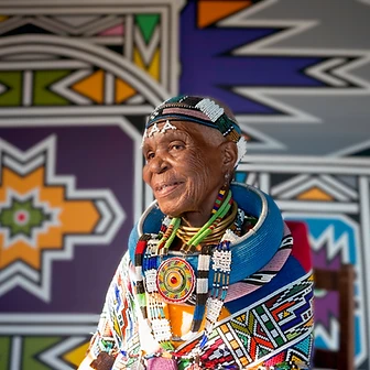 Three-quarter seated portrait of Esther Mahlangu before a mural, by Clint Strydom