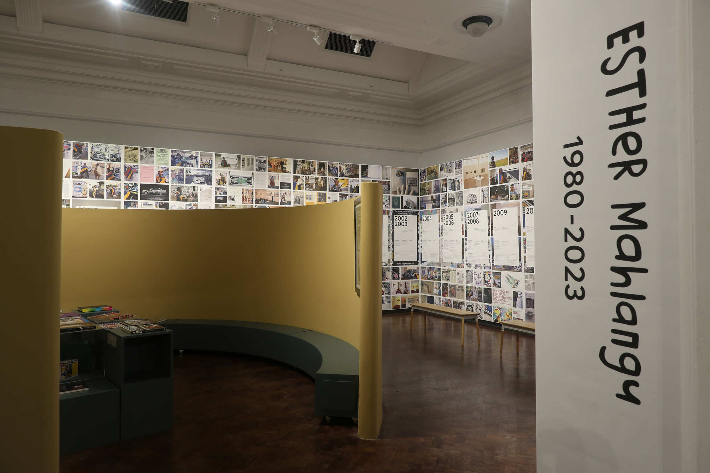 Installation view of the exhibition 'Esther Mahlangu 1980-2023' at the Iziko Museums of South Africa, with the exhibition title on a white wall and a chronological photo collage behind a mustard-yellow partition