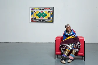 Esther Mahlangu seated in a red chair with a signed 2018 painting behind, by Clint Strydom