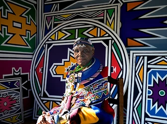 Seated portrait of Esther Mahlangu before painted Ndebele panels, by Clint Strydom, 2018