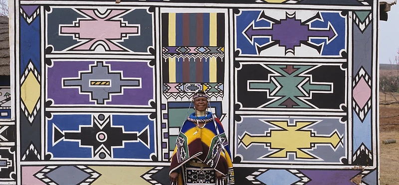 Esther Mahlangu before a completed Ndebele mural wall at Botshabelo