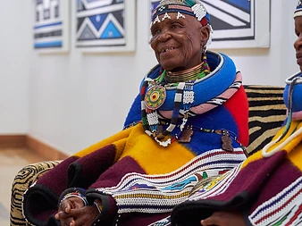Dr Esther Mahlangu seated in profile before her Ndebele canvases in the gallery