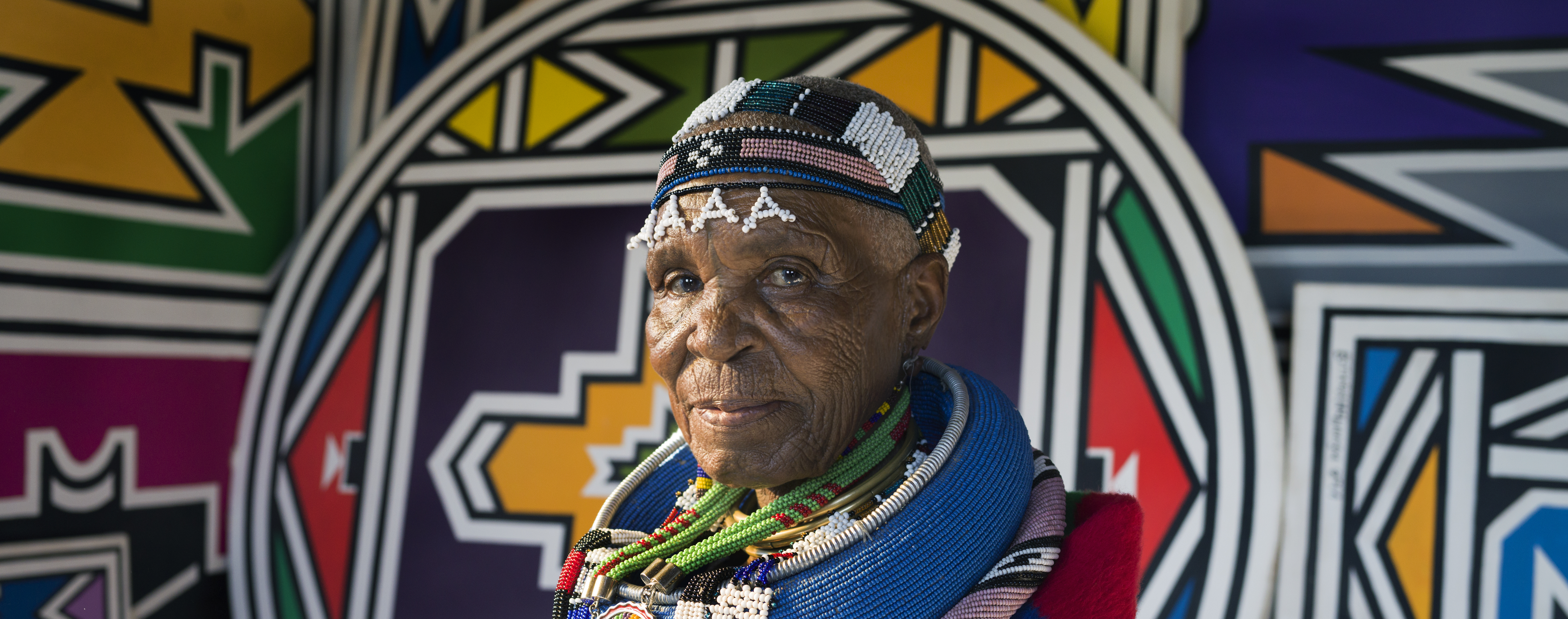 Chest-up portrait of Dr Esther Mahlangu before a painted Ndebele wall