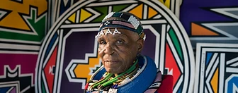 Chest-up portrait of Esther Mahlangu before a painted Ndebele wall, by Clint Strydom