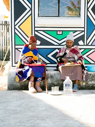 Two women sharing a meal on a step against a painted Ndebele wall, by Clint Strydom