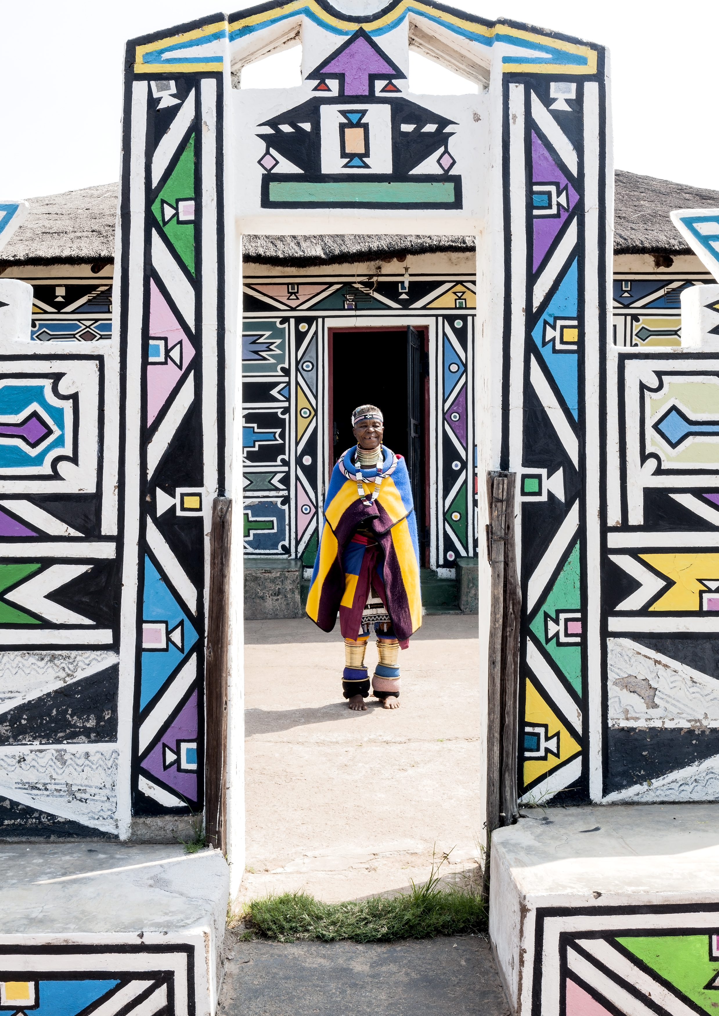 Dr Esther Mahlangu standing in the doorway of a traditional Ndebele structure, framed by hand-painted geometric walls, portrait by Clint Strydom