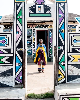 Dr Esther Mahlangu standing in the doorway of a traditional Ndebele structure, framed by hand-painted geometric walls, portrait by Clint Strydom