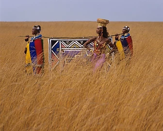Iman and two Ndebele women holding a painted canvas panel in open grassland, 1991