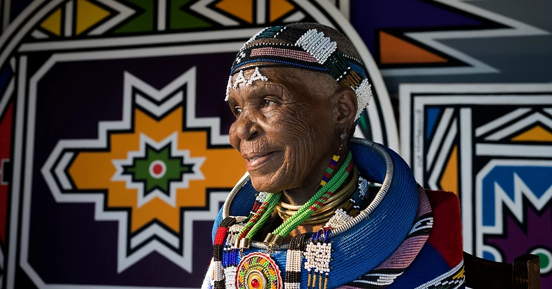 Dr Esther Mahlangu in traditional Ndebele beadwork and garment before a geometric mural — portrait by Clint Strydom